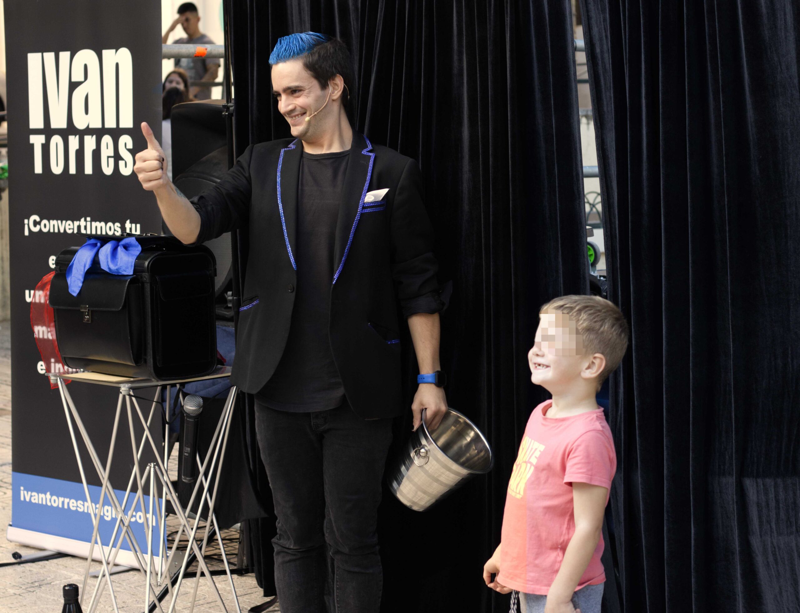 Foto de un momento del show de magia de Ivan Torres que aparece diciendo Okay con la mano con un niño al lado vestido con una camiseta rosa y sonriendo.