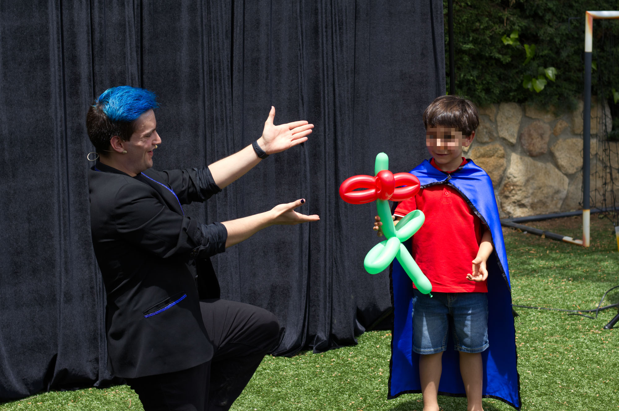 Mago cumpleaños Ivan Torres señalando al niño de cumpleaños que va con una capa azul y sujeta una flor hecha con globos