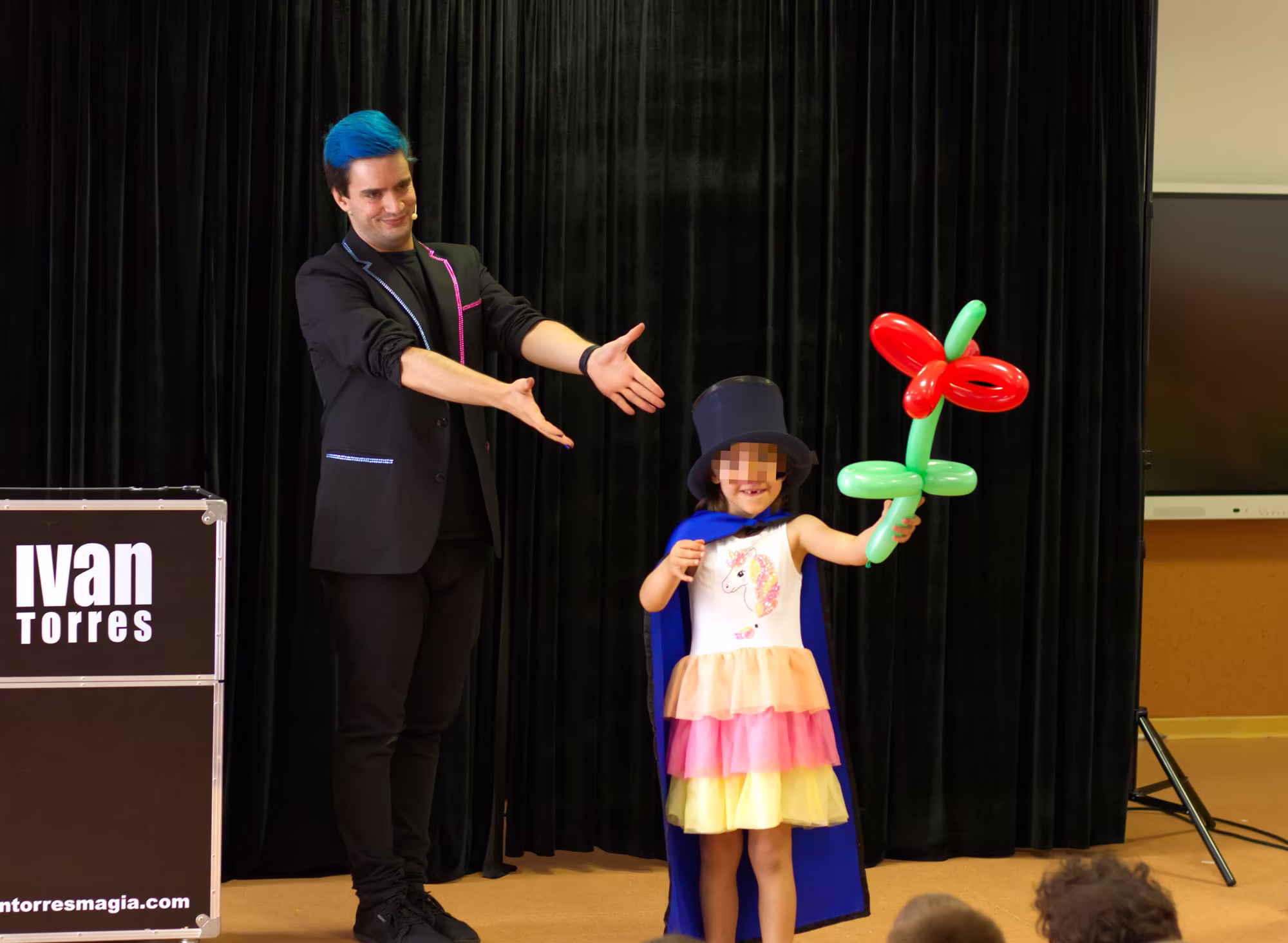 Ivan Torres Mago para colegios señalando a la niña que sujeta una flor hecha con globoflexia sonriendo